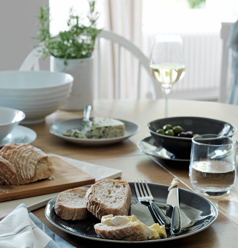 Grey and white tableware and elegant glassware are set with plates laden with rustic bread and olives.