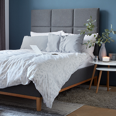 A grey bed frame, layered with white and grey bedding, sit in front of a dark blue wall.