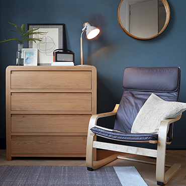 A oak vaneer chest of drawers and a sleek wood and leather chair sit infront of a deep blue accent wall.