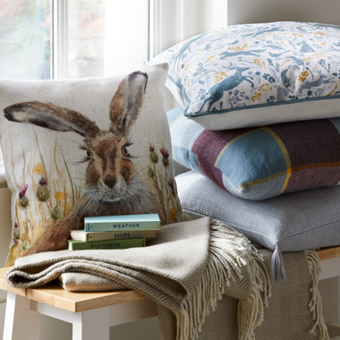 A collection of autumnal coloured check and hare print cushions sit piled-up on a window seat.