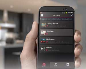 Image of a mobile phone in someone's hand, the HEOS app is displayed on the phone screen with a list of different rooms in the house.