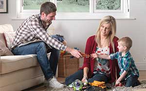 Image of a young boy sitting on the floor next to his mum and his dad sitting on the sofa passing something to him.