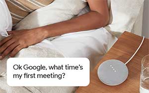 Image of someones arm whos resting in bed with a Google Mini on the bedside table. Quote asking 'Ok Google, what time is my first meeting?'