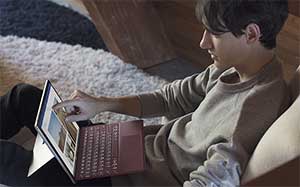 Image of a young man sitting in a lounge looking at the screen of a Microsoft Surface which is in his lap.